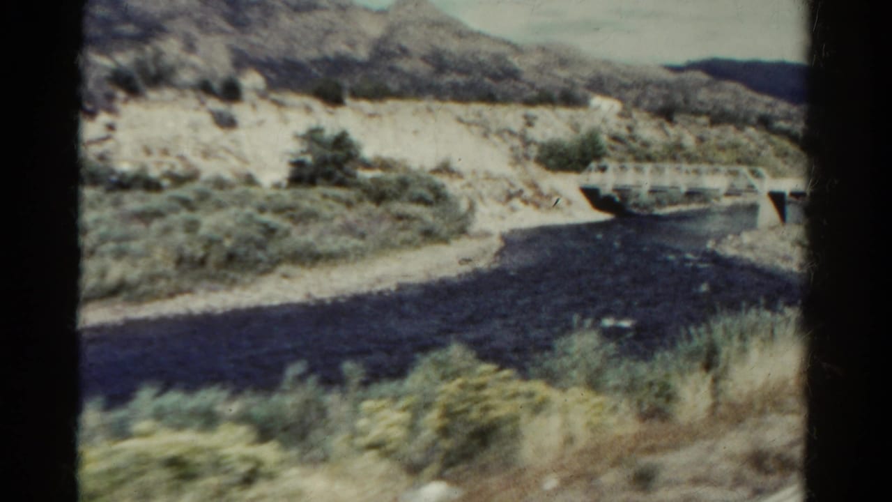 1967 Colorado 8mm frame showing a dark river, small white bridge, scrub vegetation, and pale rocky slopes in a mountain setting.