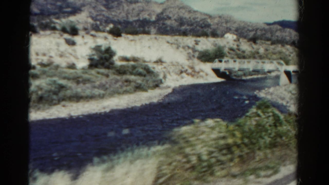 1967 Colorado home-movie frame with a river bend, white bridge, dry banks, scrub plants, and rugged hills under a bright sky.