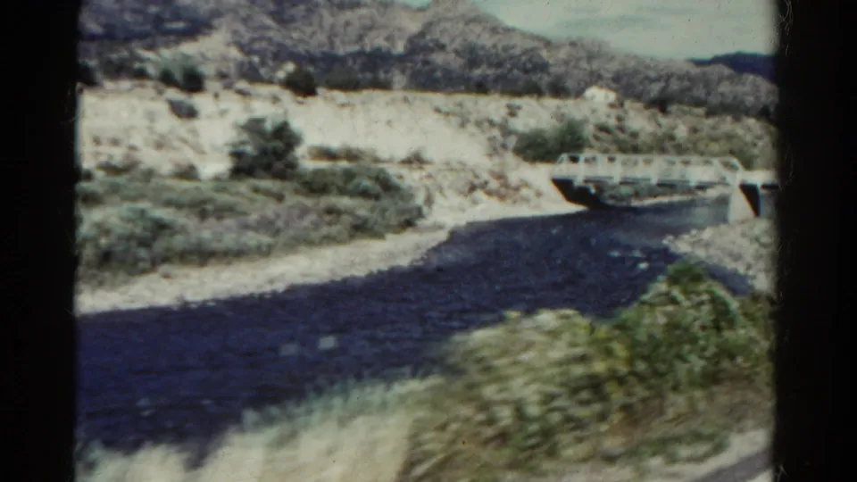 1967 Colorado home-movie frame with a river bend, white bridge, dry banks, scrub plants, and rugged hills under a bright sky.