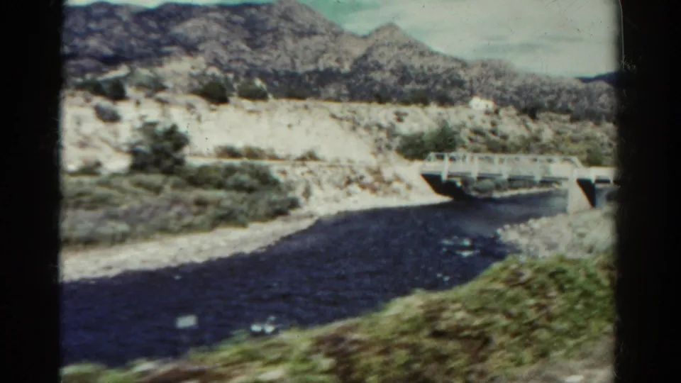 1967 Colorado 8mm still showing a white bridge crossing dark water, with dry brush, pale rocks, and mountain slopes in the background.