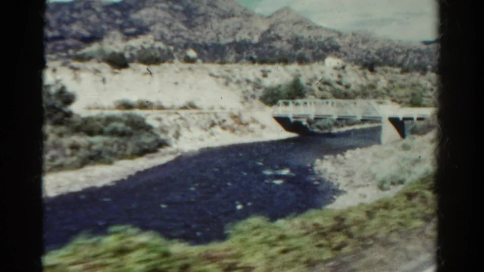 1967 Colorado frame with dark river water, a small white bridge, rocky banks, scrub vegetation, and blurred foreground greenery.