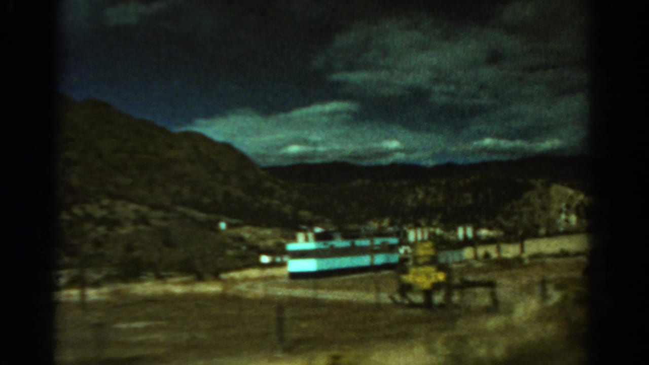 1967 Colorado 8mm frame showing a mountain slope, cloudy sky, road, and long light-blue village building.