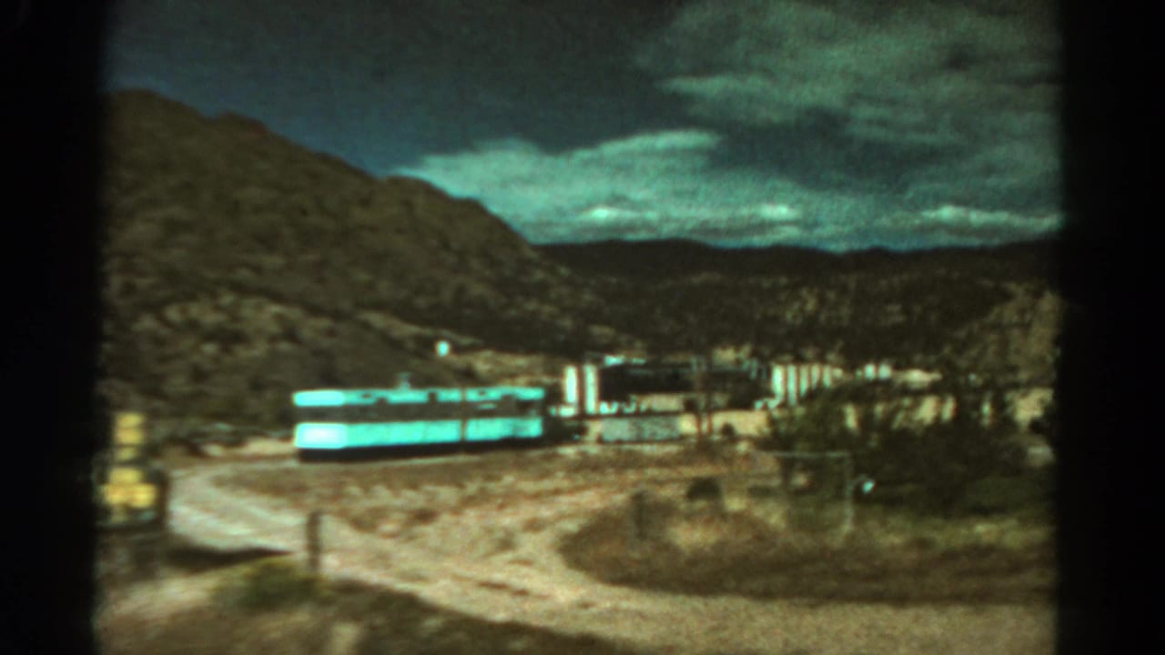 1967 Colorado home-movie frame with roadside buildings, a pale road, mountains, and heavy clouds.