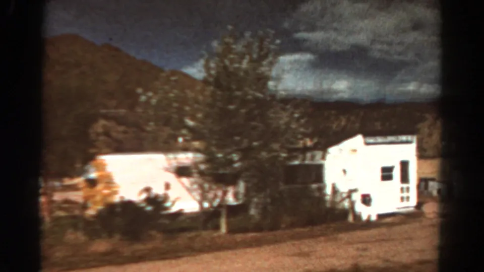 1967 Colorado frame showing a white building near a tree with mountains and clouds behind it.