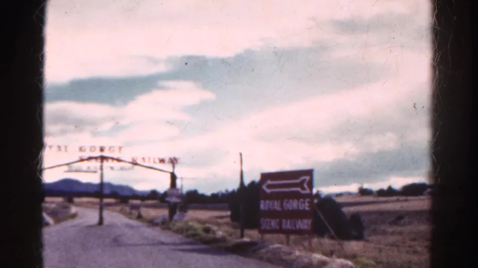 1967 Colorado home-movie view of the Royal Gorge Scenic Railway entrance arch, roadside sign, rural road, fields, hills, and cloudy sky.