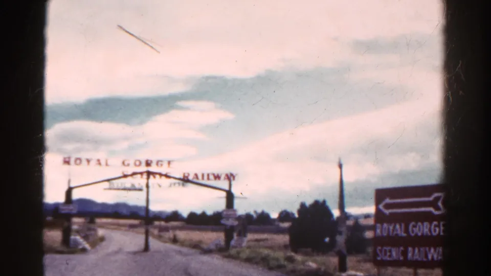 1967 Colorado 8mm frame showing the Royal Gorge Scenic Railway arch, arrow sign, road, field, distant hills, and overcast sky.