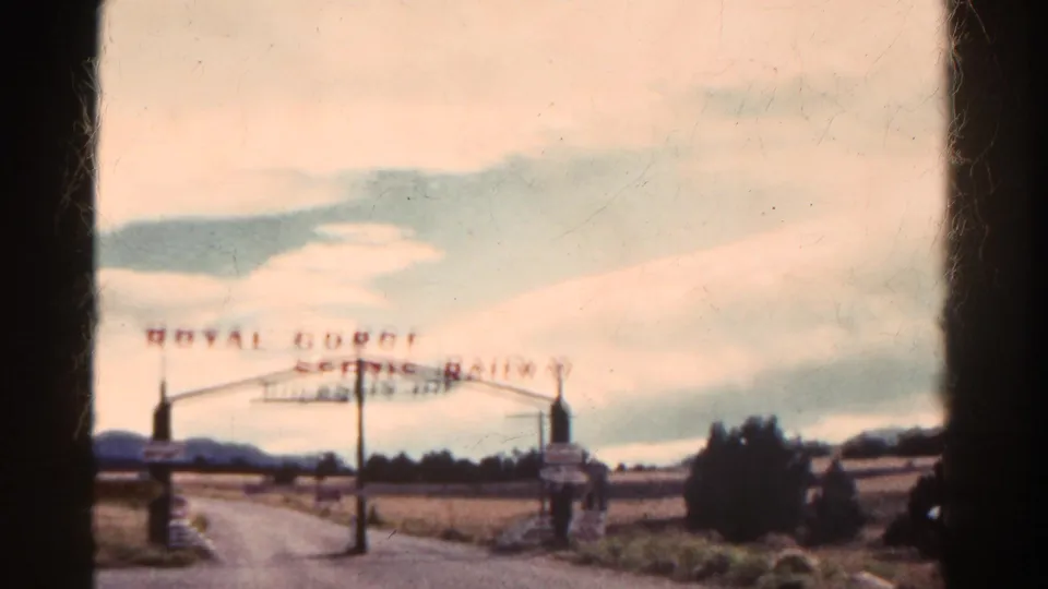 Faded 8mm view of the Royal Gorge Scenic Railway entrance in Colorado, with an arched sign, rural road, fields, and distant hills.