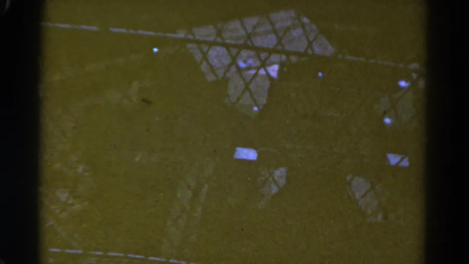 Dark 1967 Colorado 8mm frame showing chain-link fencing, greenish shadow, and faint distant structural or landscape shapes.