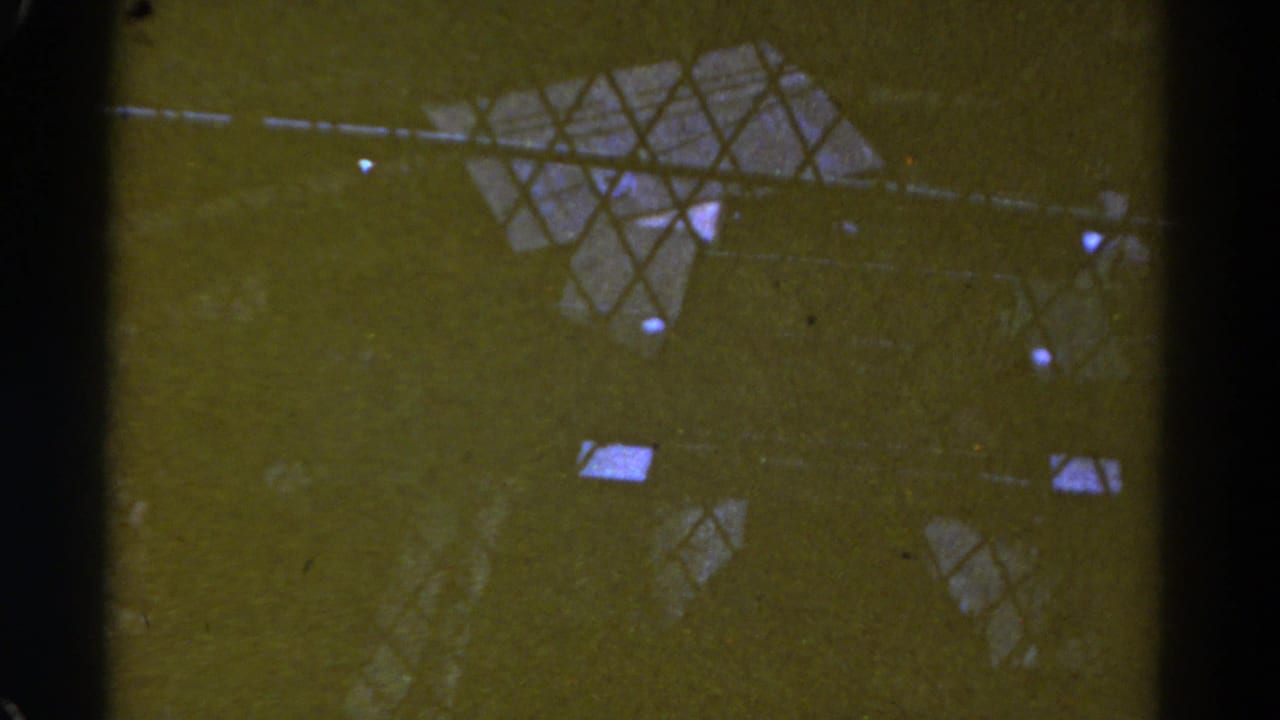 1967 Colorado 8mm frame showing dark vertical bars, diamond-pattern mesh, and small bright reflections across a green-yellow industrial surface.