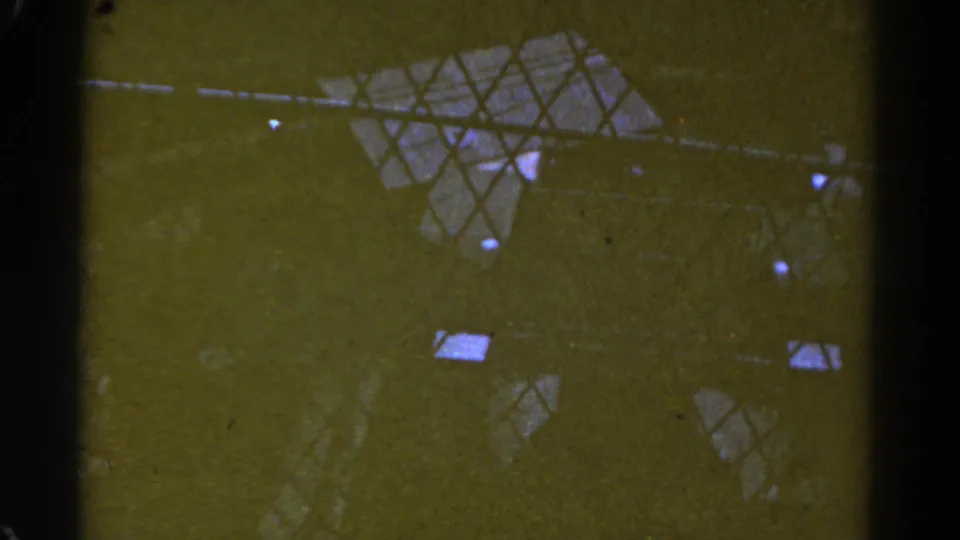 1967 Colorado 8mm frame showing dark vertical bars, diamond-pattern mesh, and small bright reflections across a green-yellow industrial surface.