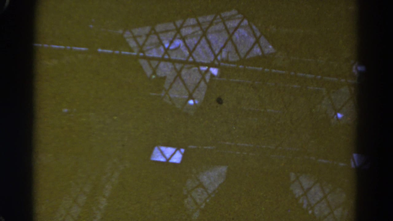 1967 Colorado 8mm frame with wire mesh, dark vertical supports, and scattered light reflections on a greenish industrial surface.