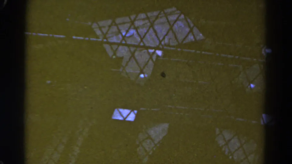 1967 Colorado 8mm frame with wire mesh, dark vertical supports, and scattered light reflections on a greenish industrial surface.