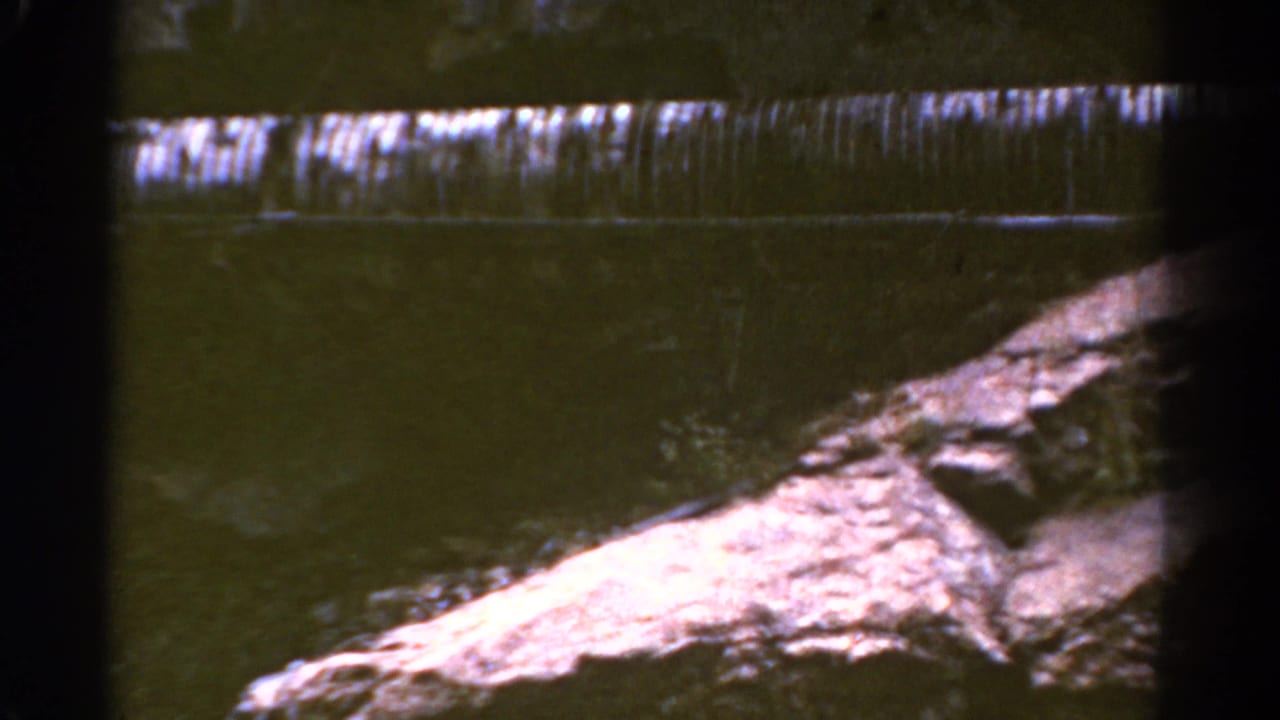 1967 Colorado 8mm frame showing water falling over a low dam or ledge beside rocks and greenery.