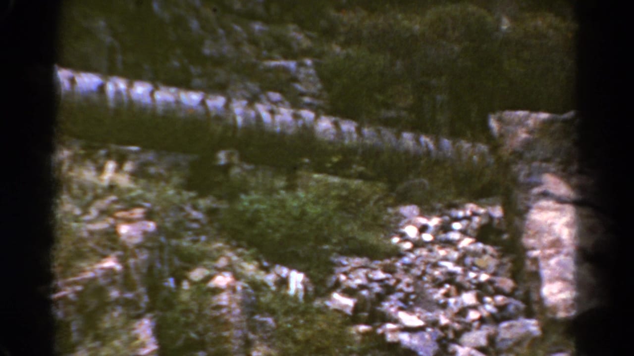 1967 Colorado 8mm frame of a rugged streamside area with rocks, brush, and shallow water.