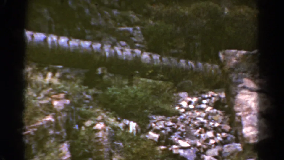 1967 Colorado 8mm frame of a rugged streamside area with rocks, brush, and shallow water.