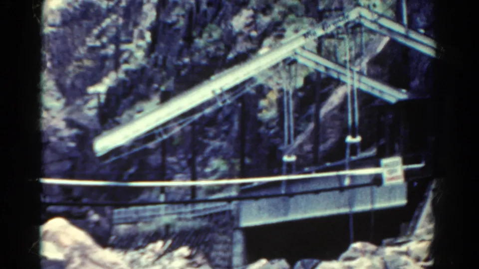 1967 Colorado home-movie frame showing a dam or flume structure with railings and signs in a canyon.