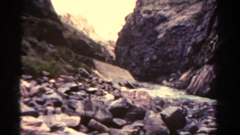 1967 Colorado canyon view with a stony riverbed, large foreground boulders, water, and bright sun on distant rock walls.