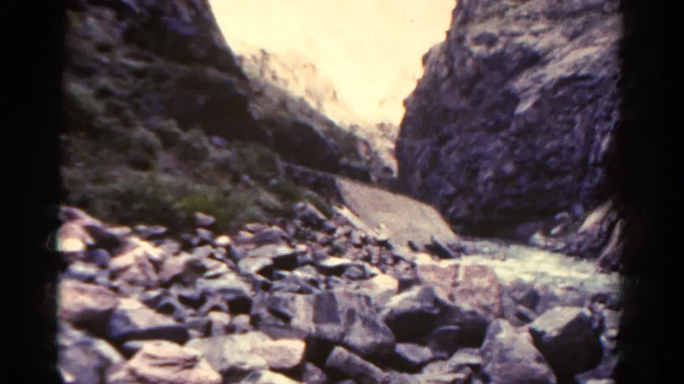 Colorado 8mm home-movie frame looking up a rugged canyon, with scattered rocks, a shallow stream, and sunlit cliffs beyond.