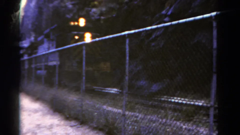 1967 Colorado 8mm frame with a dark train moving past a chain-link fence beside canyon rock.