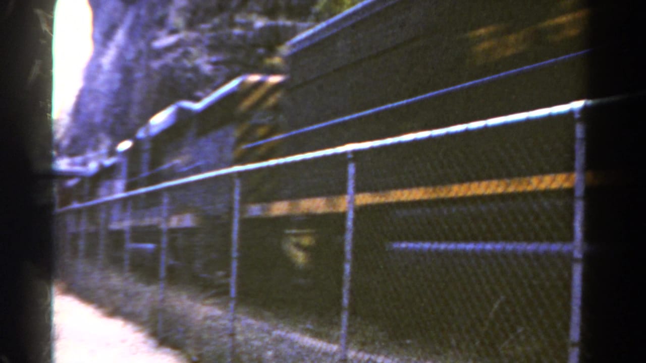 1967 Colorado 8mm frame showing a dark freight train moving past a chain-link fence along a rocky rail corridor.