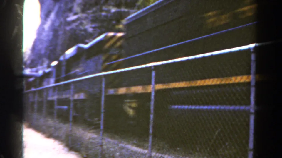 1967 Colorado 8mm frame showing a dark freight train moving past a chain-link fence along a rocky rail corridor.