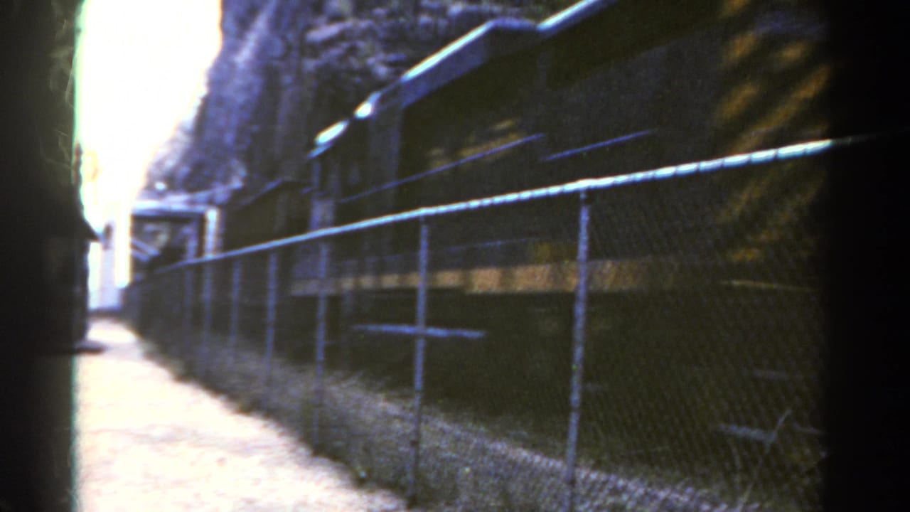 Train cars pass behind a fence with a bright opening ahead.