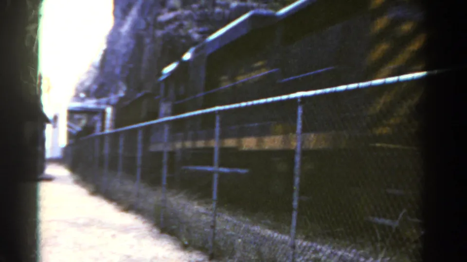 1967 Colorado 8mm view of freight cars moving through a fenced corridor beside rock, with bright exposure ahead.
