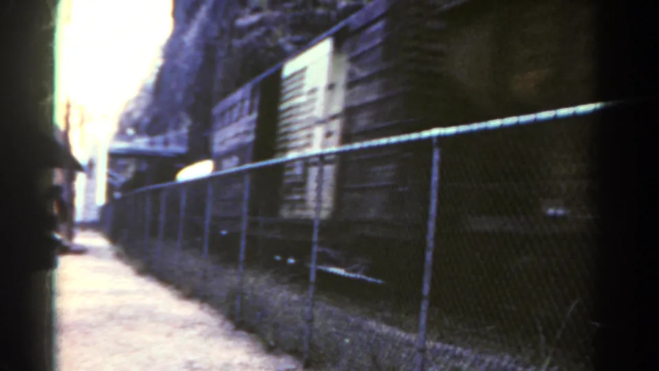 1967 Colorado 8mm frame with a freight car passing behind chain-link fencing along a narrow trackside path.