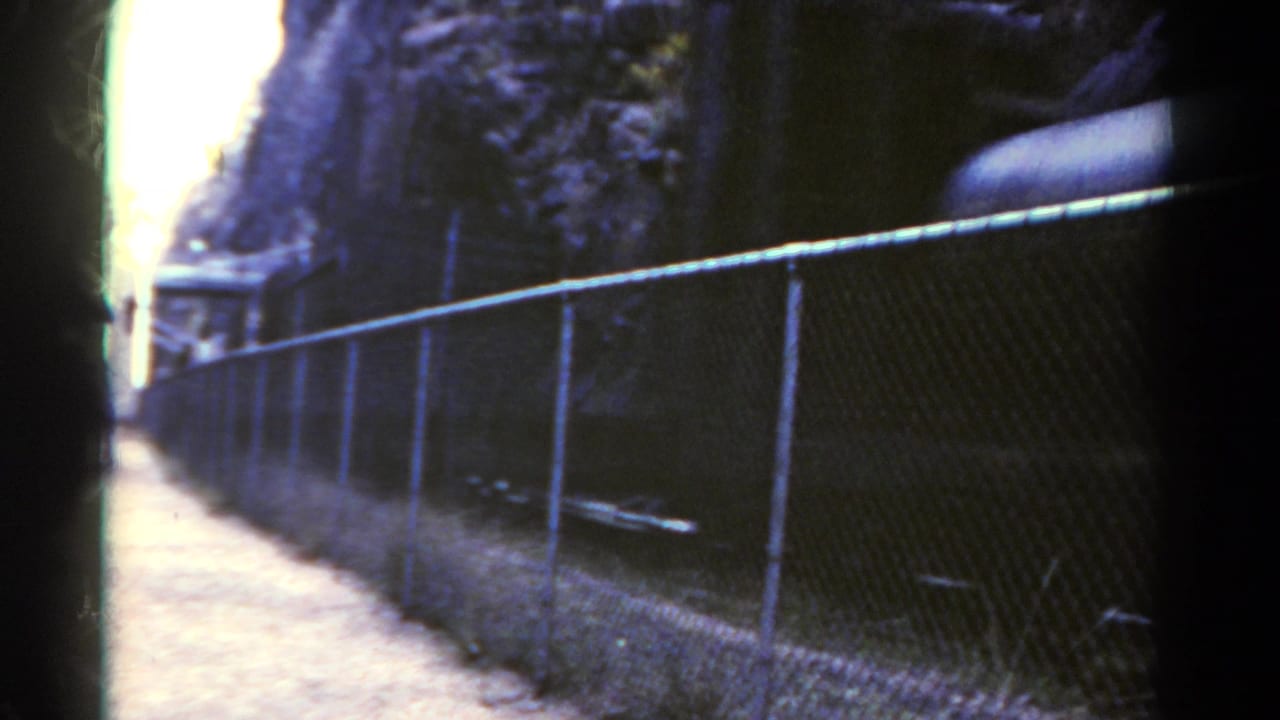 1967 Colorado 8mm frame showing a chain-link fence along a dirt path beside a mountain rail corridor before the freight cars fill the view.
