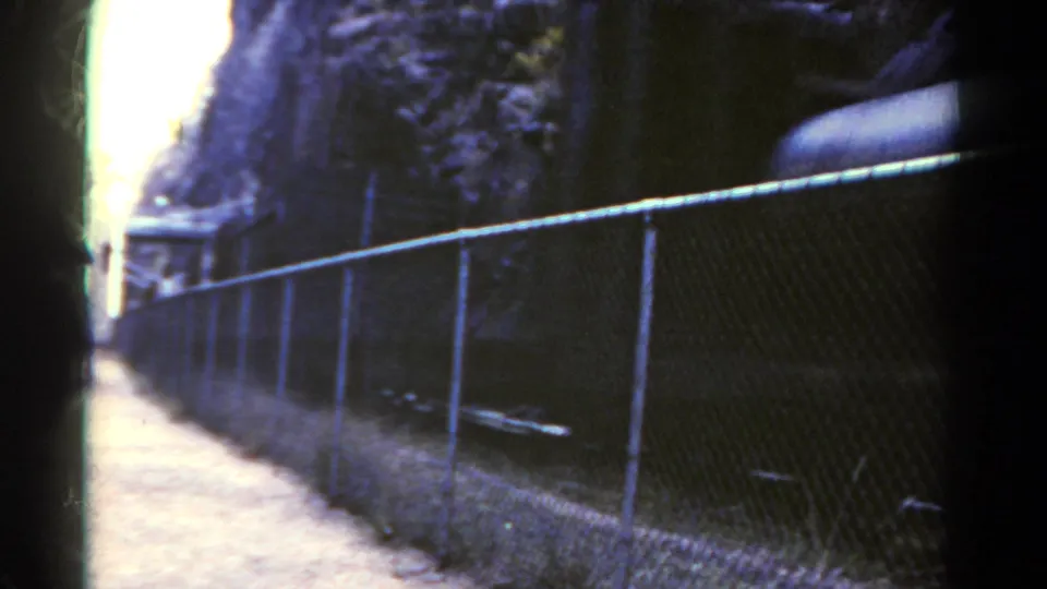 1967 Colorado 8mm frame showing a chain-link fence along a dirt path beside a mountain rail corridor before the freight cars fill the view.