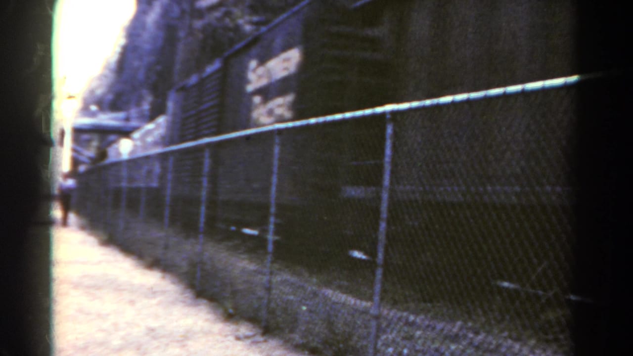 Fence and dirt path beside a dark rail corridor with a small distant figure.