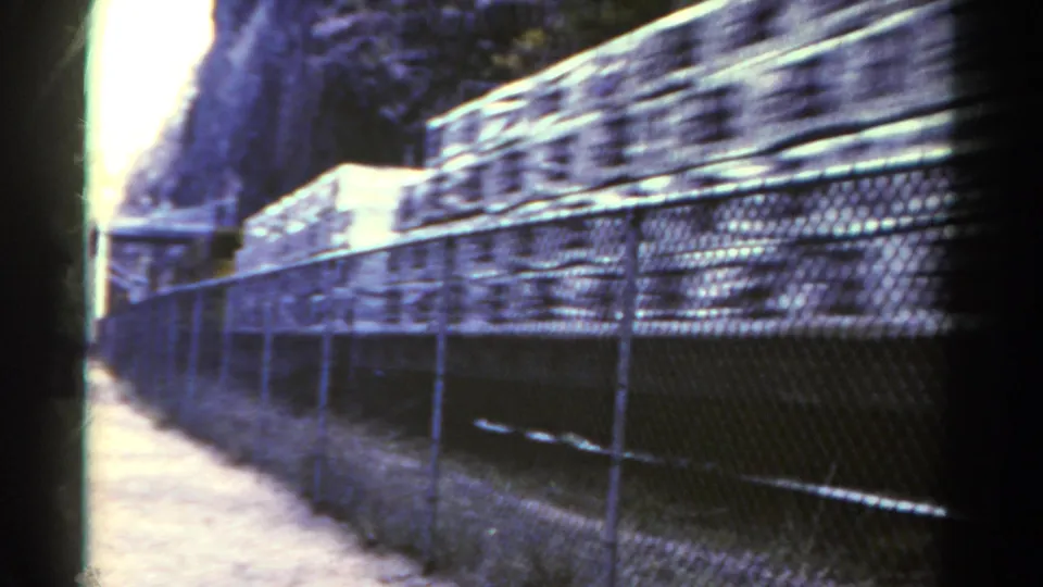 1967 Colorado 8mm frame showing freight cars rushing behind a chain-link fence beside a dirt path in a mountain rail corridor.