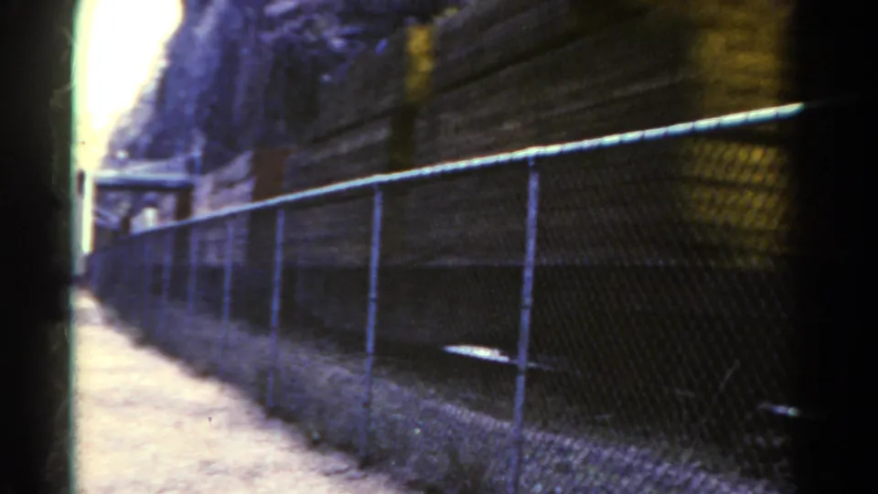 1967 Colorado home-movie frame with the train moving out of view behind a chain-link fence, with path, slope, and rail corridor still visible.
