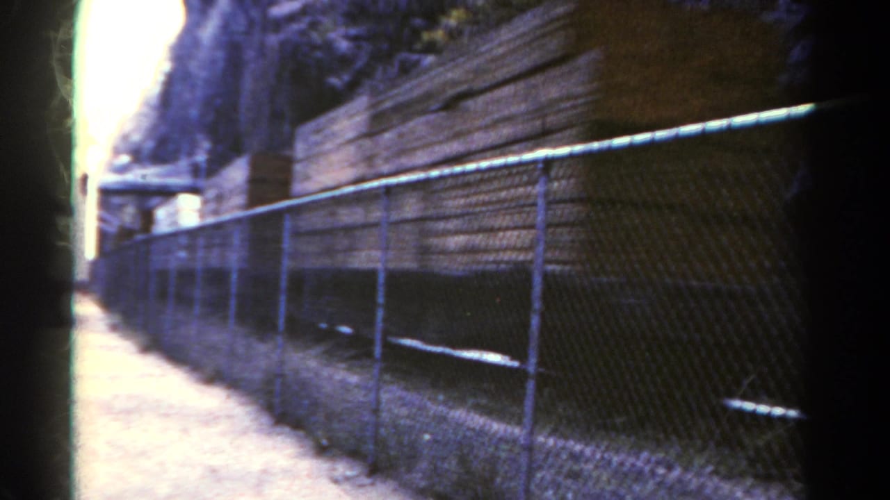 1967 Colorado 8mm frame showing a chain-link fence along a rail-side dirt path with stacked materials and a steep rocky backdrop.