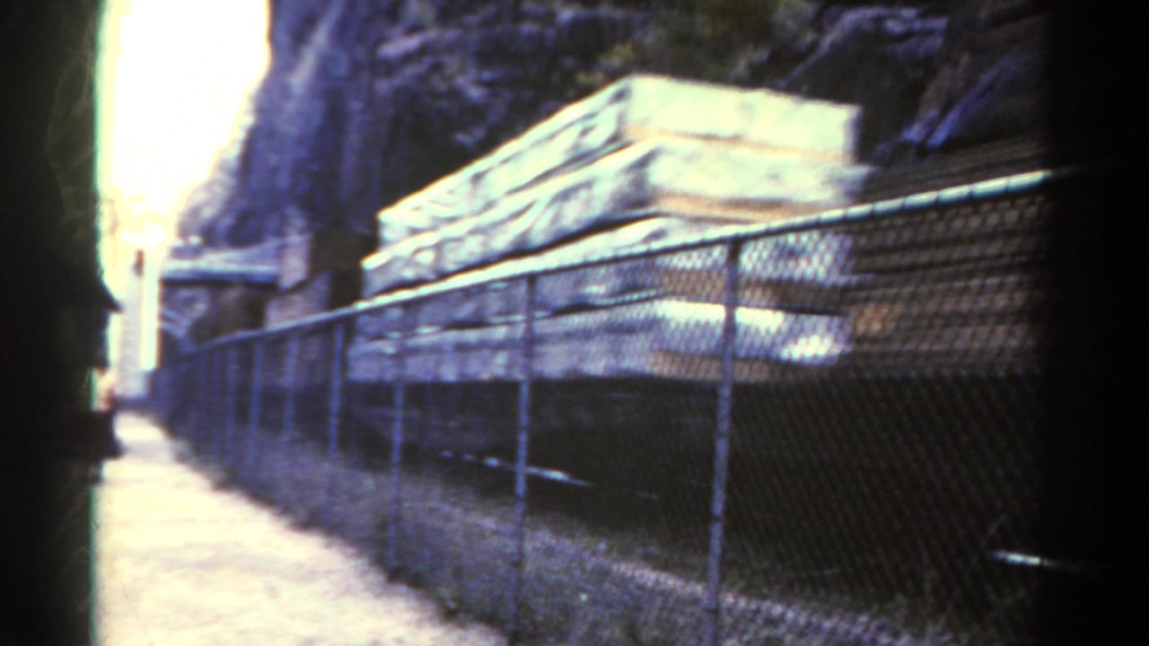Fence-lined path with pale stacked materials beside a rocky rail corridor.