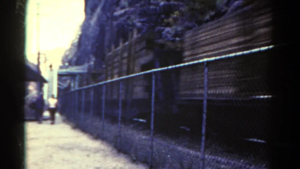 1967 Colorado 8mm frame showing a fence-lined dirt path, utility poles, and small distant people near a mountain rail corridor.