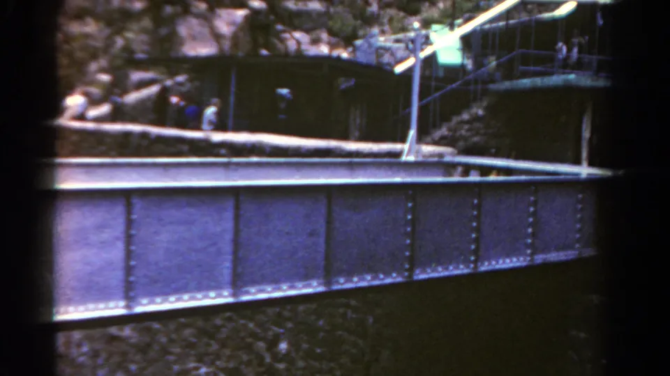 1967 Colorado frame showing a riveted metal bridge or rail structure with railings, rocky terrain, and small distant people above it.