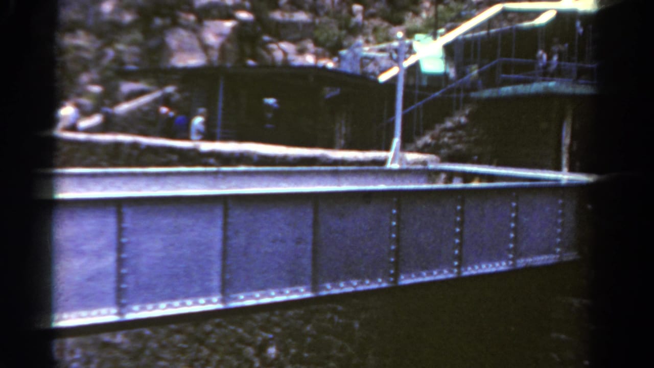 1967 Colorado 8mm frame showing a riveted bridge span below a rocky overlook, with railings, a small structure, and distant visitors.