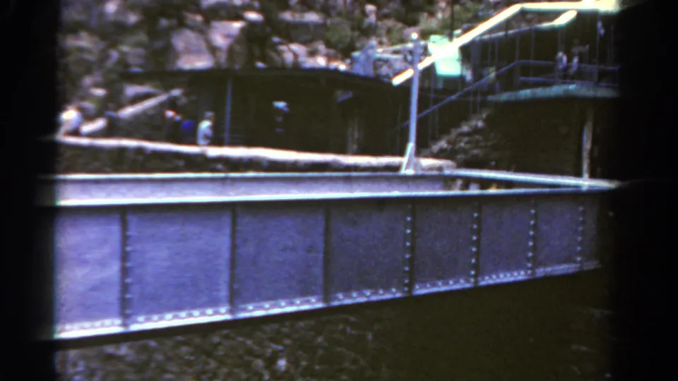 1967 Colorado 8mm frame showing a riveted bridge span below a rocky overlook, with railings, a small structure, and distant visitors.