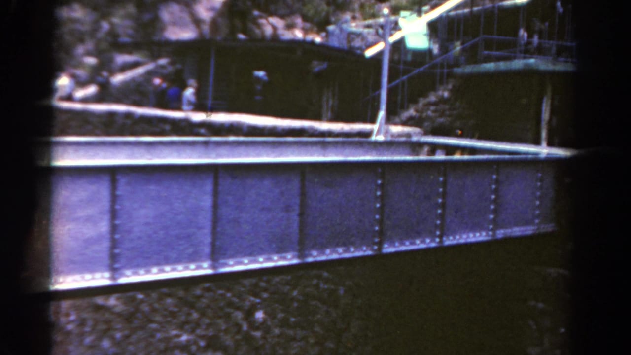 1967 Colorado home-movie frame with a riveted bridge in front of a rocky canyon overlook, stairway, railings, and distant people.