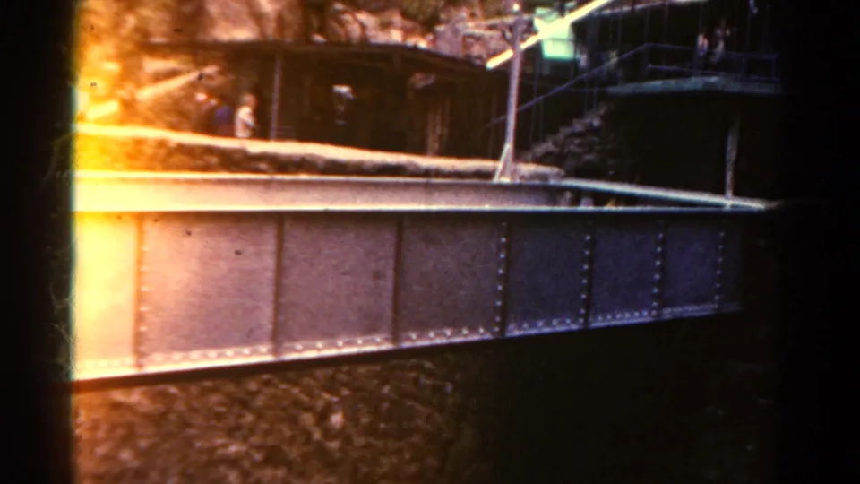 Warm-shifted 1967 Colorado 8mm frame showing the riveted bridge, canyon overlook, stairs, railings, and distant visitors.
