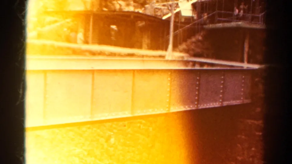 1967 Colorado 8mm frame with heavy orange light leak across a riveted bridge, rocky canyon, railings, and faint distant figures.
