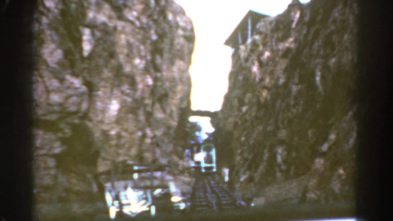 Small train entering a narrow rocky canyon with tracks and a cliffside structure visible.