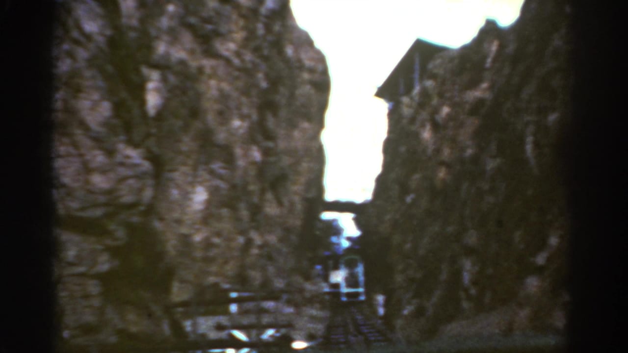 Rocky canyon view with a small train near the lower center and a building high on the right.