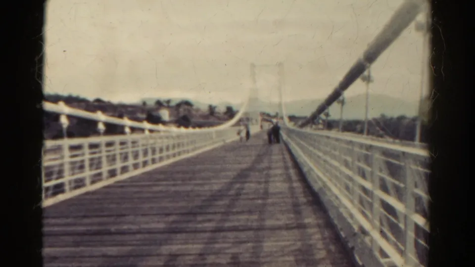 1967 Colorado travel frame showing a wooden pedestrian suspension bridge with white railings and distant visitors crossing.