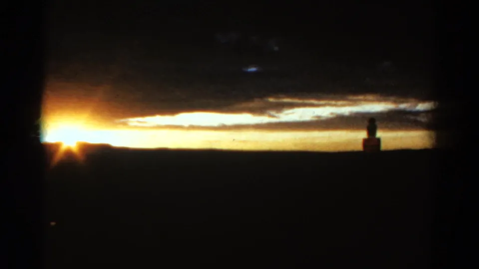 Vintage Colorado 8mm sunset frame with the sun near the horizon, dark clouds overhead, and a small distant silhouetted structure or marker.