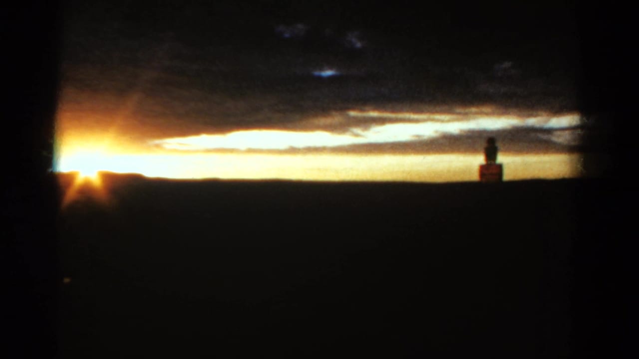 Low sun on a dark horizon with clouds and a small silhouetted figure near a distant structure.