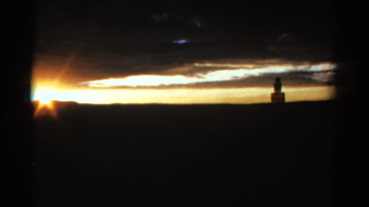 1967 Colorado home-movie frame with a golden sunset, layered clouds, dark horizon, and a distant person silhouetted near a small structure.