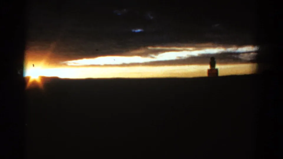 1967 Colorado 8mm frame showing intense sun flare, a dark country horizon, cloud layers, and a small silhouetted figure near the right edge.