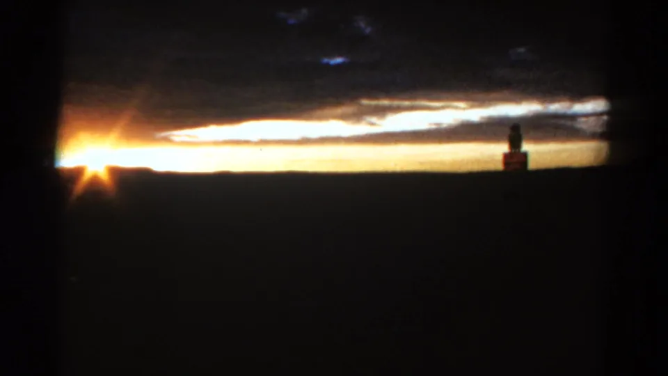 Final 1967 Colorado sunset frame with warm sky, dark rural horizon, clouds, and a distant standing figure beside a small silhouetted structure.